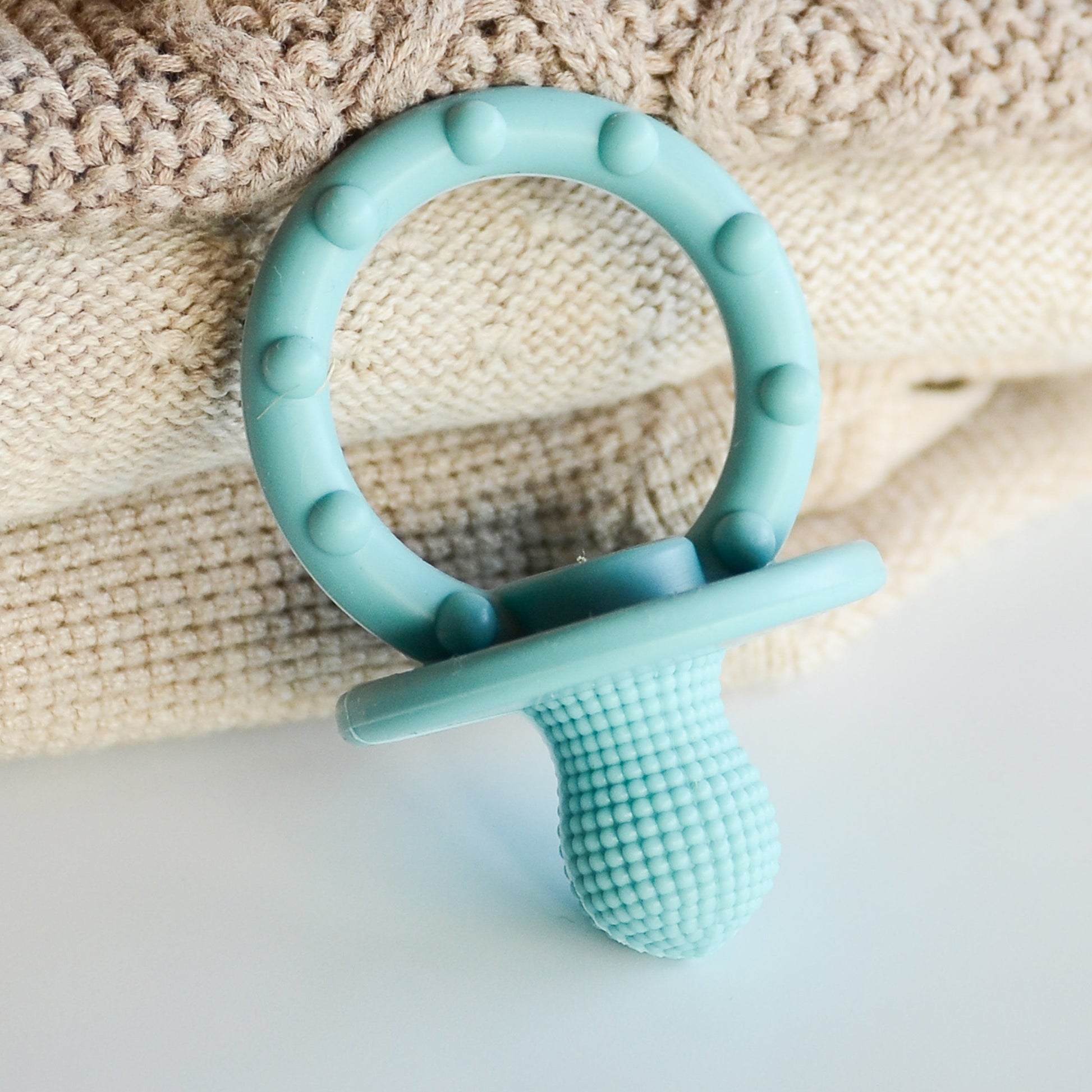 A light blue silicone baby pacifier teether with a textured shield and a circular ring, resting on a beige knitted blanket over a white surface.