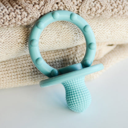 A light blue silicone baby pacifier teether with a textured shield and a circular ring, resting on a beige knitted blanket over a white surface.