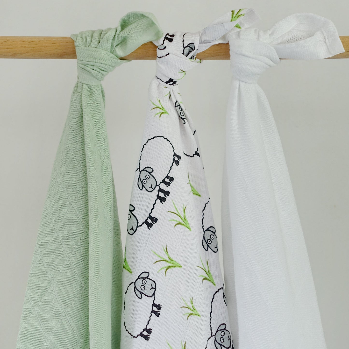 Two baby swaddle blankets, one green and one with sheep design, hanging on a wooden rod against a white background.