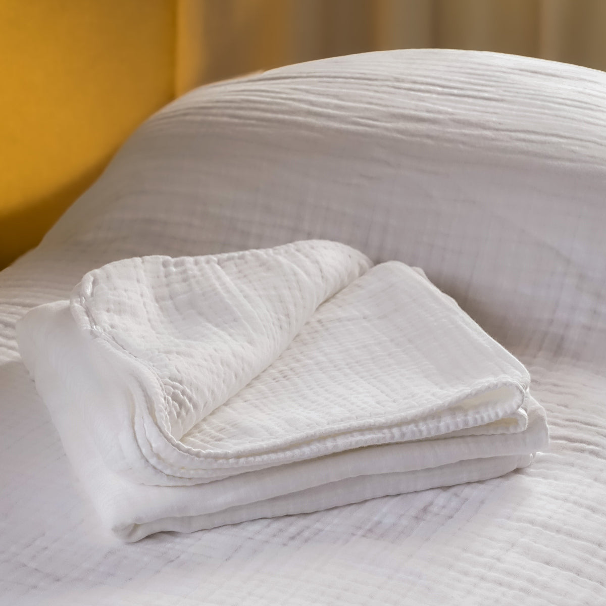 A folded white textured muslin blanket or cotton sheet rests on a bed with matching white bedding and a warm yellow headboard in the background.