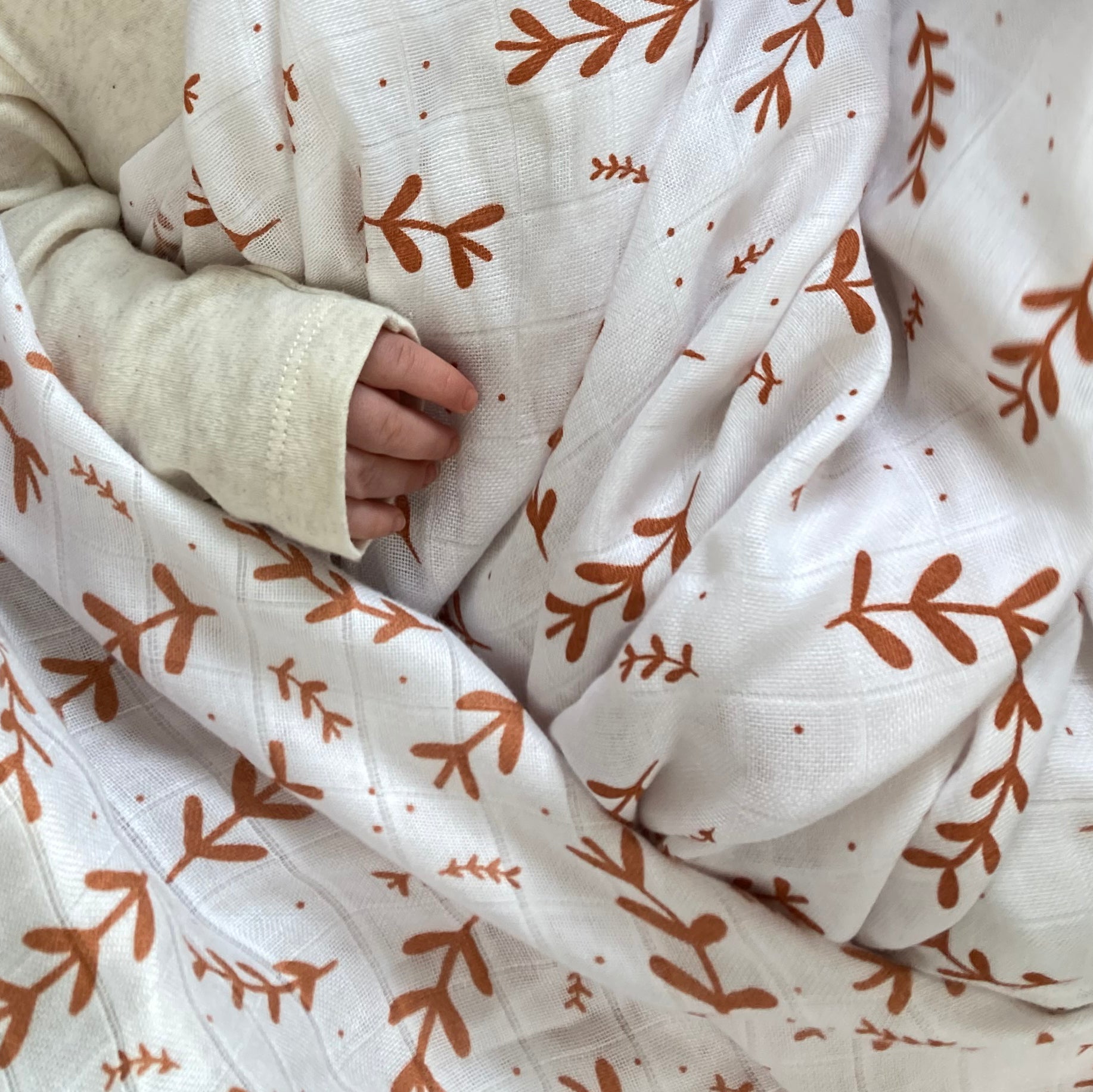Close-up of a baby wrapped in a white blanket with brown leaf patterns, wearing beige pants.